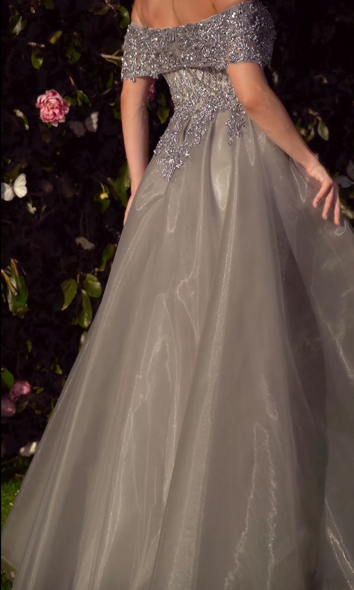 Wearing the Andrea & Leo MA103 Long Silver Prom Ball Gown, a person stands with their back to the camera, showcasing beaded details and a mermaid silhouette against a dark floral backdrop.